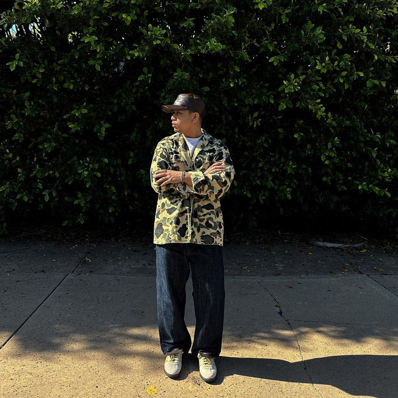 @26mm.jpeg wearing a camo chore coat, wide leg dark wash denim pants, and a leather baseball cap @26mm.jpeg wearing a camo chore coat, wide leg dark wash denim pants, and a leather baseball cap