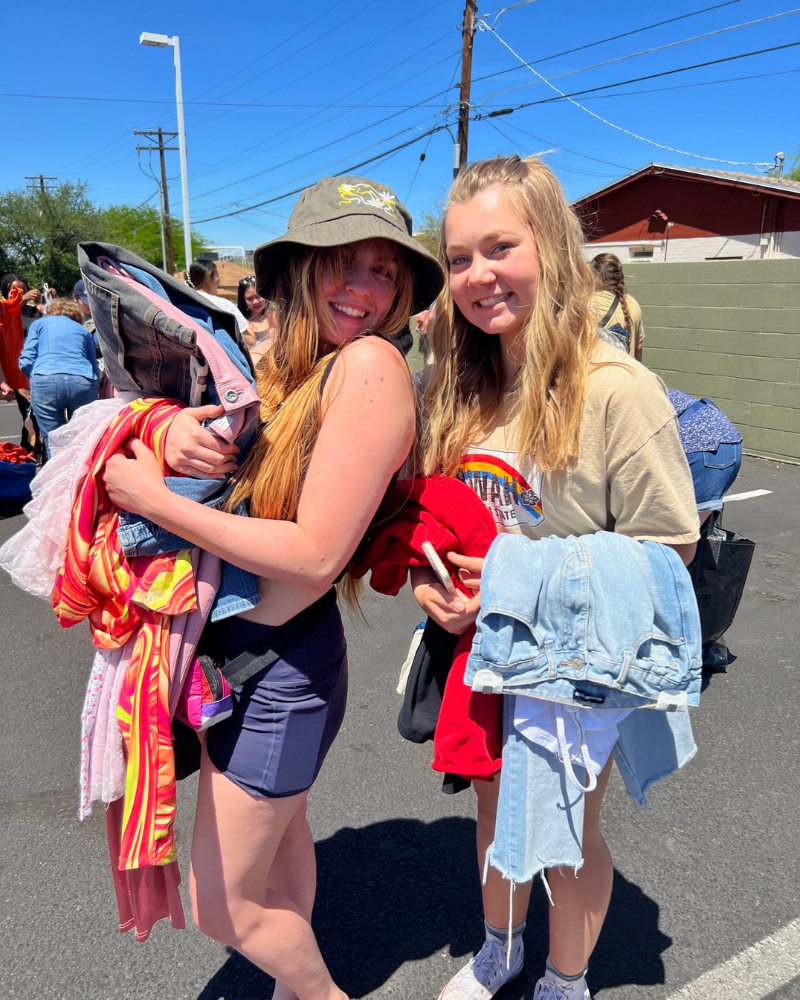 two friends smile at camera while holding clothing in a parking lot
