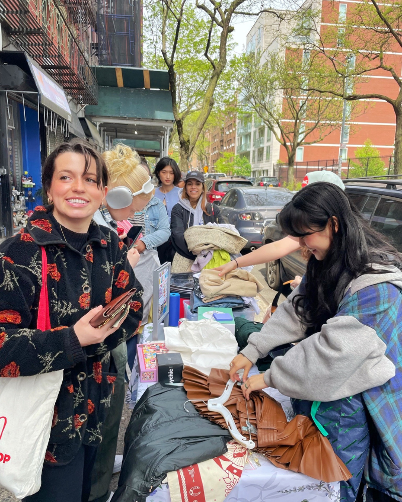 person faces the camera and smiles while other shoppers go through clothes set on table