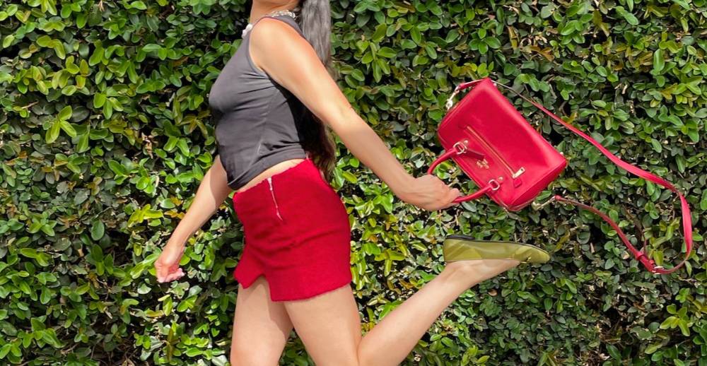 Person in a black tank top, red mini skirt, and olive ballet flats running down a sidewalk with a Coach bag in hand