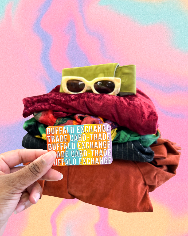 Hand holding a Buffalo Exchange trade card in front of a stack of clothing and accessories